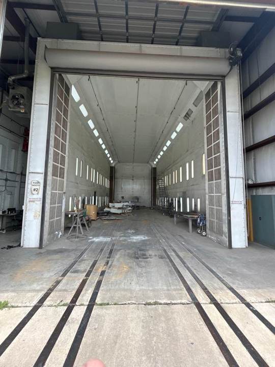 Drive-through industrial paint booth with tall doors open and rail tracks in the floor